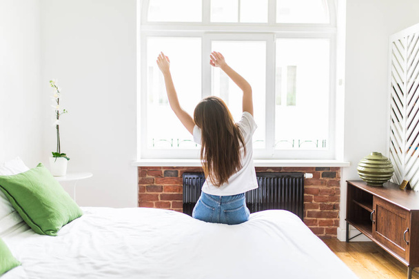 Mujer de pie en su cama cerca de la ventana mientras se estira después de despertarse con el amanecer por la mañana, vista trasera
 - Foto, Imagen
