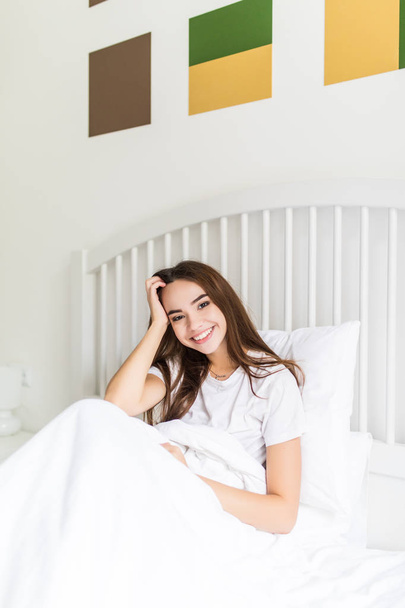 Красивая молодая женщина с подушкой сидит в постели и улыбается фотоаппарату дома
 - Фото, изображение