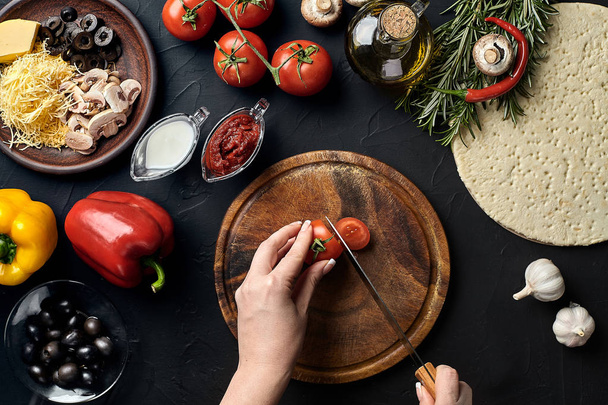 Female hand cut tomatoes on wooden board on kitchen table, around lie ingredients for pizza: vegetables, cheese and spices. - Photo, Image