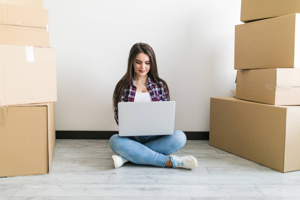 Happy young woman sitting on floor near moving boxes. Young woman moving to new home. Woman using laptop sitting on floor around cardboxes - Photo, Image