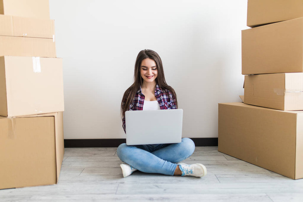 Молодая женщина переезжает в новую квартиру, сидит на полу вокруг коробки и серфинга в Интернете на ноутбуке в поисках новых идей украшения
. - Фото, изображение