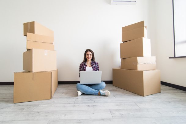 Attractive young woman is moving sitting among cardboard boxes, using a laptop in new apparment - Photo, Image