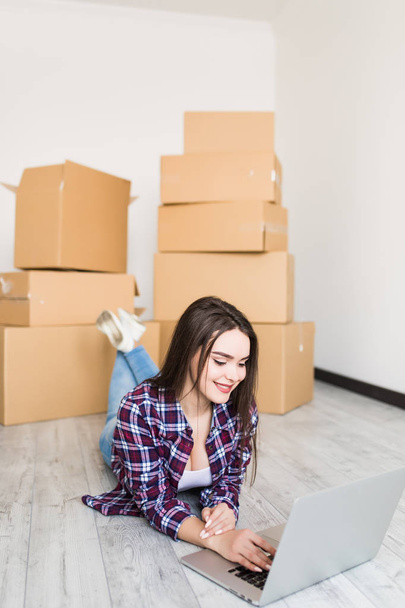 Young woman is moving using a laptop and smiling, lying among cardboard boxes in new home - Photo, Image