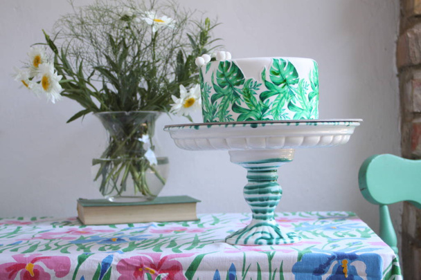 Holiday cake with palm leaves design. Tropical birthday party inspiration. Tasty cake with wafer paper monstera leaves on table with flowers in vase  - Photo, Image