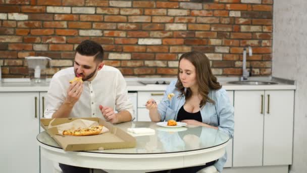 Man en zwangere vrouw eten pizza thuis in hun keuken. Slechte voeding. Vette voedingsmiddelen. Obesitas - Video