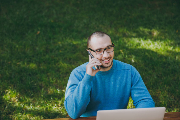 Young successful smiling smart man businessman or student in casual blue shirt, glasses sitting at table, talking on mobile phone in city park using laptop, working outdoors. Mobile Office concept. - Photo, Image