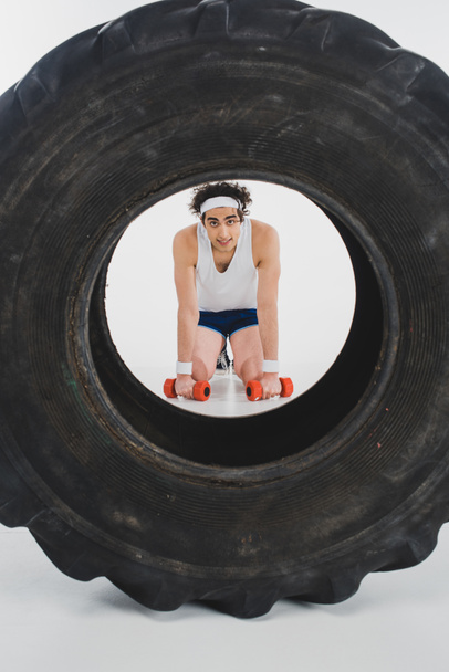 junger dünner Sportler sitzt mit Hanteln in der Hand mit Reifen im Vordergrund - Foto, Bild