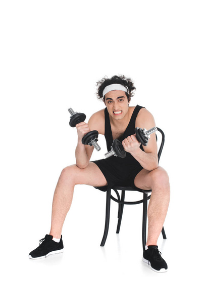 Sandalye üzerinde oturan beyaz izole iken dumbbells ile egzersiz genç sıska sporcu - Fotoğraf, Görsel