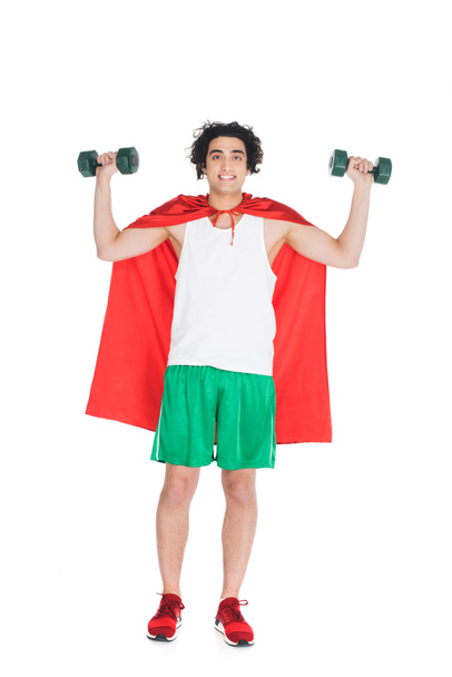 Smiling skinny sportsman with dumbbells standing in red cape isolated on white - Zdjęcie, obraz