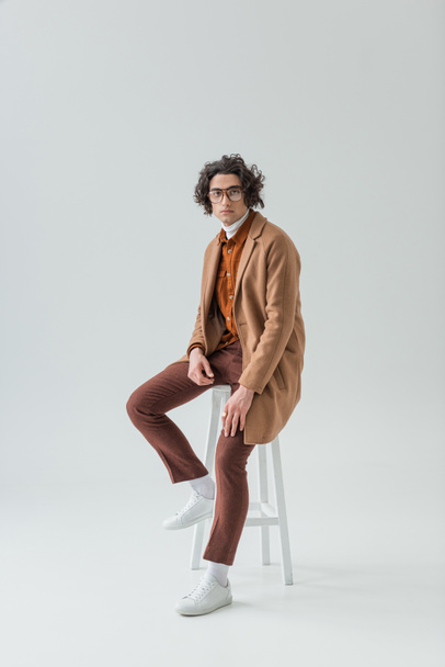 Young stylish man in eyeglasses sitting on chair isolated on grey - Foto, imagen