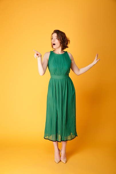 joven sorprendida mujer en vestido verde apuntando lejos aislado en naranja
 - Foto, Imagen