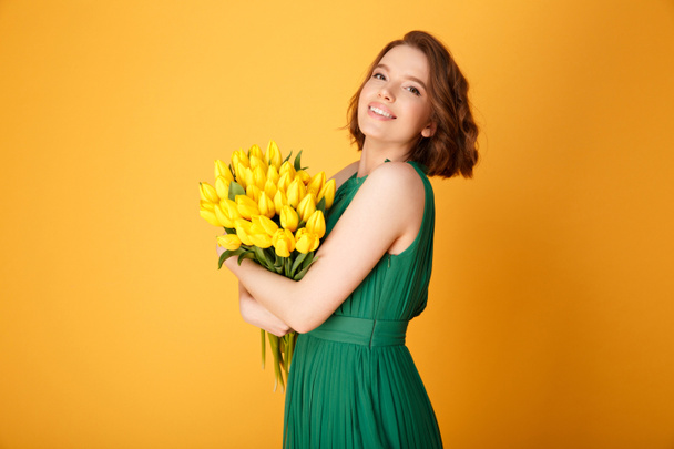 side view of beautiful woman with bouquet of yellow tulips looking at camera isolated on orange - Photo, Image