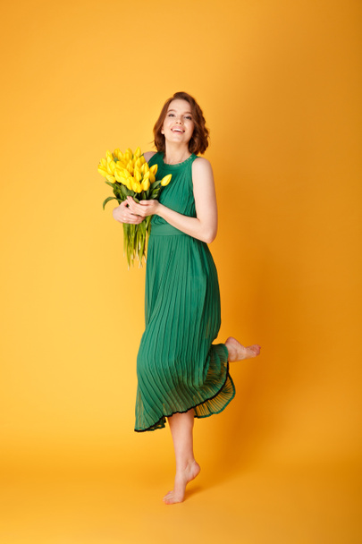 mooie jonge vrouw in groene jurk met boeket geel voorjaar tulpen geïsoleerd op oranje - Foto, afbeelding