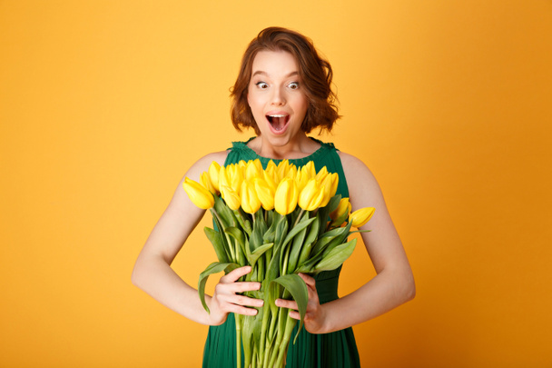 retrato de mujer sorprendida mirando ramo de tulipanes amarillos en manos aisladas en naranja
 - Foto, imagen