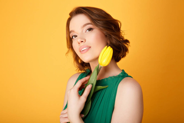 portrait of attractive woman in green dress with yellow tulip isolated on orange - Foto, Bild
