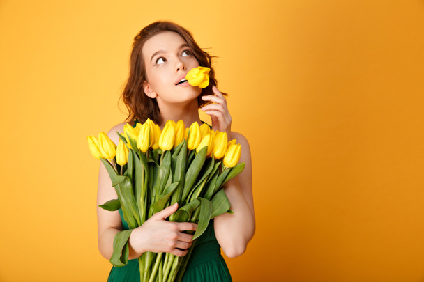 portrait of pensive woman with tulip in mouth and bouquet of yellow tulips isolated on orange - Valokuva, kuva