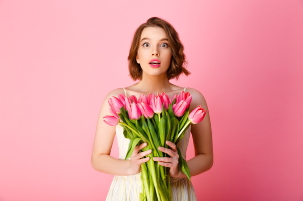 döbbenve, fiatal nő, látszó-on fényképezőgép, elszigetelt rózsaszín rózsaszín tulipán csokor - Fotó, kép