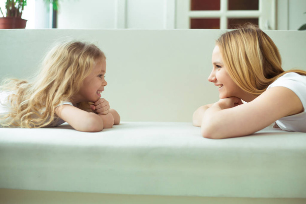 Moms and daughters. Young beautiful woman and a little blond girl lie on a white sofa face to face and look at each other. - 写真・画像