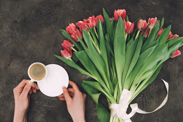 partial top view of person holding cup of coffee with saucer and beautiful tulip flowers - Zdjęcie, obraz