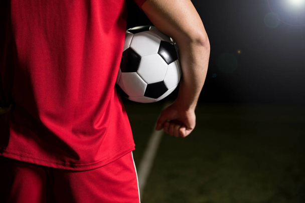 Viljelty peruutusnäkymä nuori jalkapalloilija punainen univormu pitämällä palloa kainalossaan stadionilla
 - Valokuva, kuva