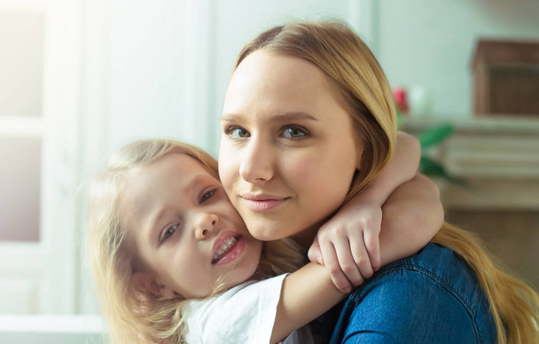 Beautiful smiling blond mother and daughter embrace and have fun on the couch at home and look at the camera. Concept of mother and daughter. - Photo, Image