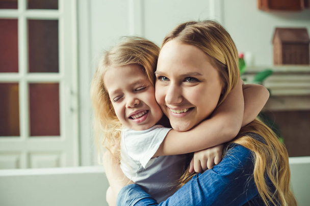 Beautiful smiling blond mother and daughter embrace and have fun on the couch at home and look at the camera. Concept of mother and daughter. - Valokuva, kuva