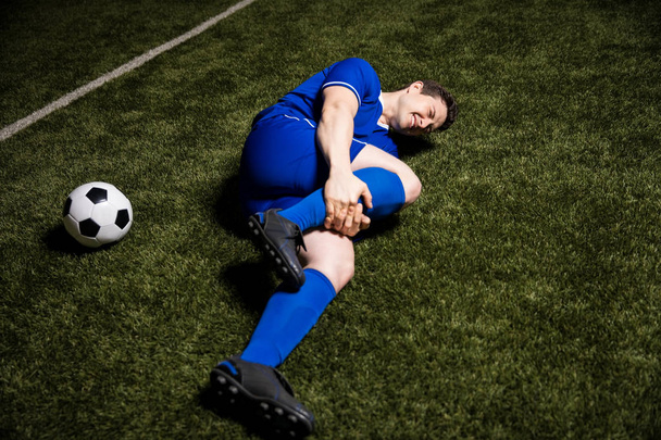 Giocatore di calcio ferito sdraiato a terra e tenendo la gamba durante una partita di calcio
 - Foto, immagini