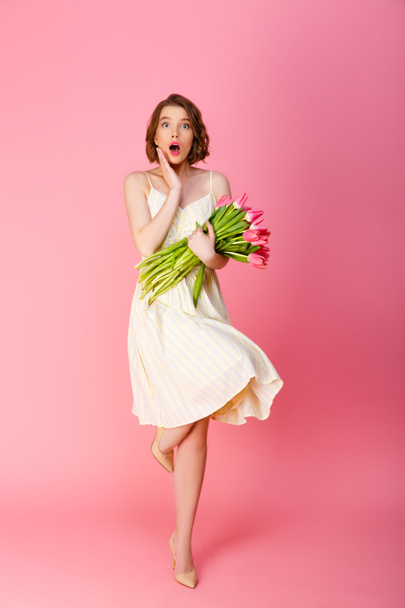 young shocked woman with bouquet of pink tulips looking at camera isolated on pink - Fotó, kép