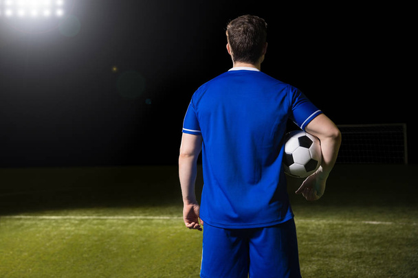Vista trasera del jugador de fútbol masculino en jersey azul con una pelota bajo el brazo en el estadio
 - Foto, imagen