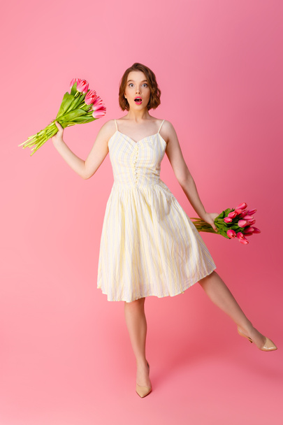 шокированная женщина в белом платье с букетами весенних цветов изолированы на розовый
 - Фото, изображение