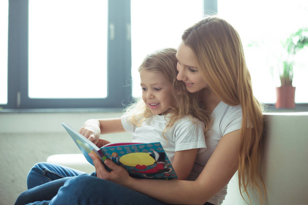 Beautiful smiling blond mother and daughter are sitting on the couch in an embrace. Mom is reading a story to her daughter or a fairy tale from a book. - Foto, Imagen