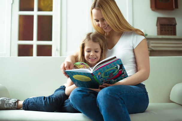 Beautiful smiling blond mother and daughter are sitting on the couch in an embrace. Mom is reading a story to her daughter or a fairy tale from a book. - Zdjęcie, obraz