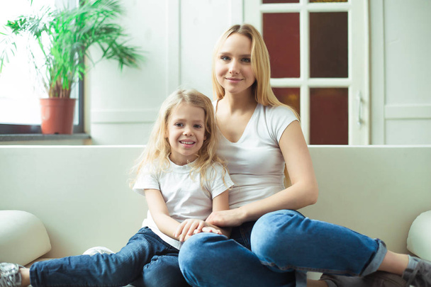 Beautiful smiling blond mother and daughter embrace and have fun on the couch at home and look at the camera. Concept of mother and daughter. - Photo, Image