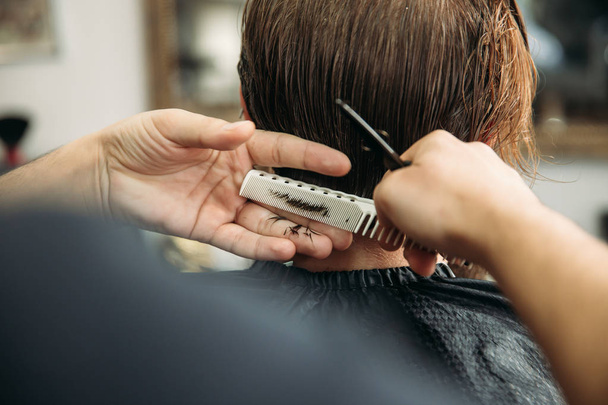 die Hände eines jungen Friseurs, der einem attraktiven bärtigen Mann im Friseursalon eine Frisur schneidet - Foto, Bild