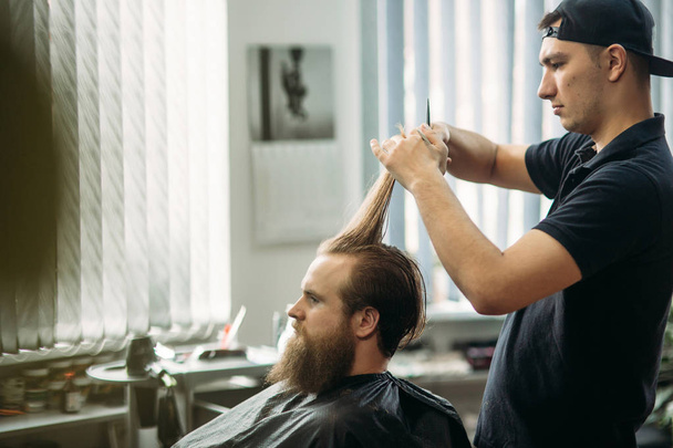 Mestre corta cabelo e barba de homens na barbearia, cabeleireiro faz penteado para um jovem
 - Foto, Imagem