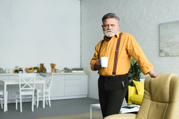 hombre mayor barbudo con estilo sosteniendo la taza de café y mirando a la cámara en casa
 - Foto, imagen