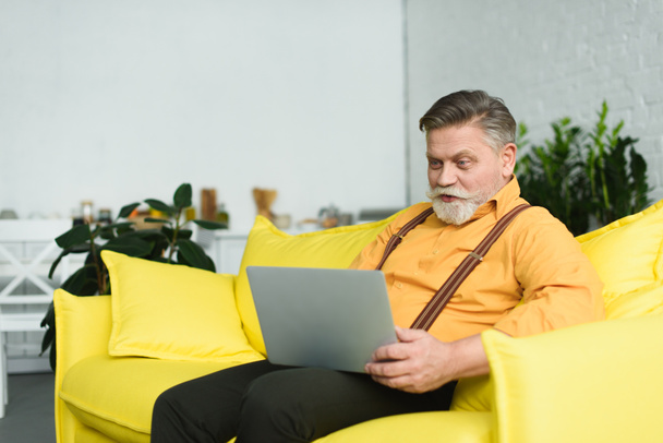 hymyilevä parrakas vanhempi mies käyttää kannettavaa tietokonetta istuessaan sohvalla kotona
 - Valokuva, kuva