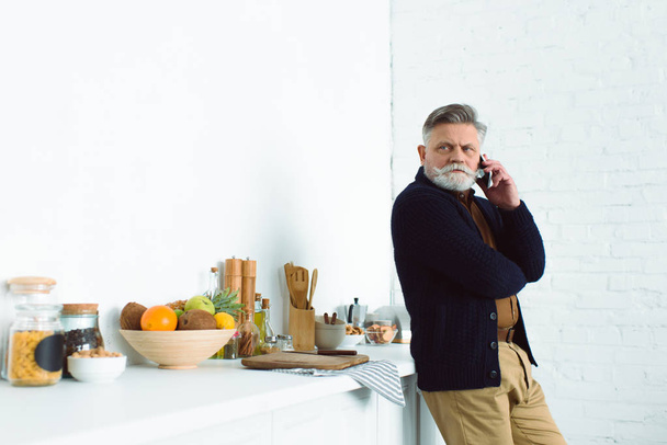 bebaarde senior man praten door de smartphone tijdens het permanent in de keuken en op zoek weg - Foto, afbeelding