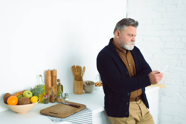 bearded senior man using smartphone while standing at kitchen  - Photo, Image