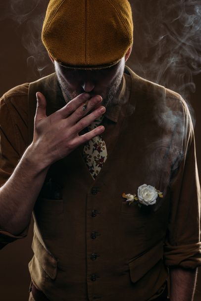 Stijlvolle man in platte dop roken sigaret geïsoleerd op brown - Foto, afbeelding