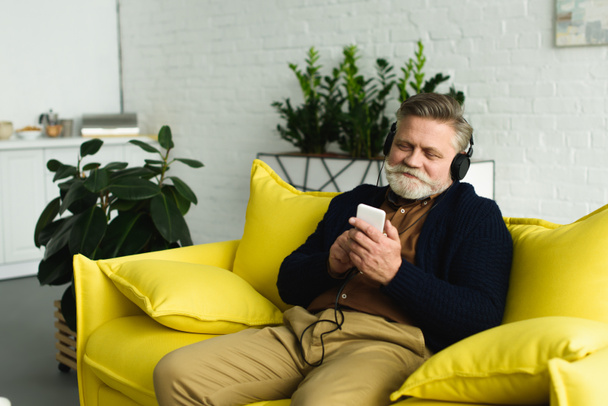 smiling bearded senior man in headphones sitting on sofa and listening music with smartphone - 写真・画像