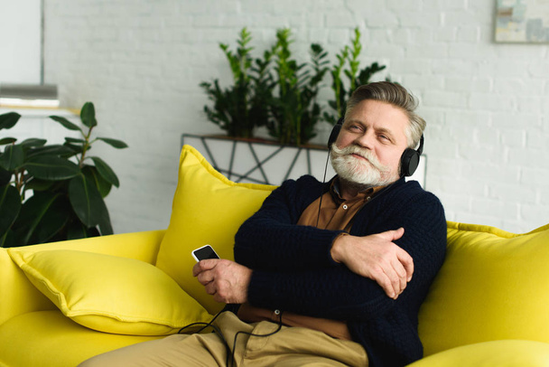 счастливый пожилой человек в наушниках слушает музыку со смартфона дома
 - Фото, изображение