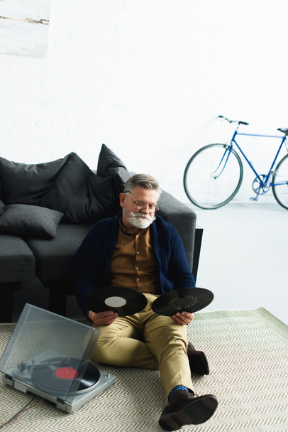 vysoký úhel pohledu vousatý starší muž v brýlích, zatímco sedí na koberci doma drží vinylových desek  - Fotografie, Obrázek