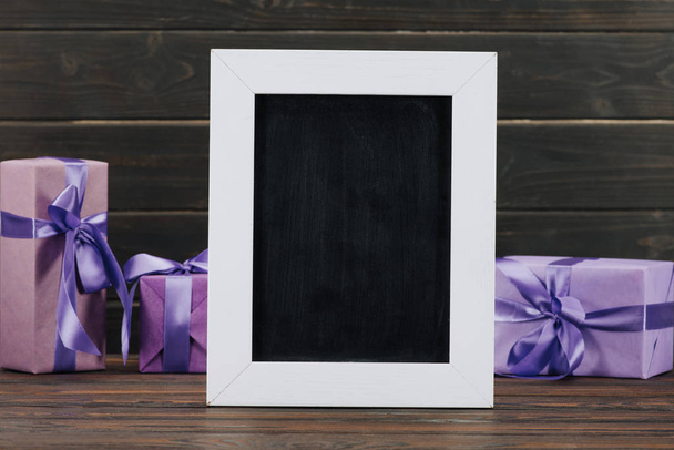 pizarra en blanco en marco con cajas de regalo contra pared de madera
 - Foto, Imagen