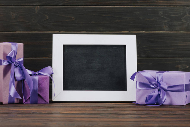 blank blackboard in frame with gift boxes against wooden wall - Фото, зображення