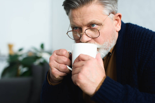 close-up view of senior man in eyeglasses drinking from cup and looking away at home - Foto, Imagem