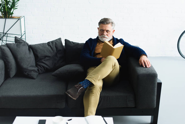 стильный бородатый мужчина в очках держит книгу и смотрит в камеру, сидя дома на диване
 - Фото, изображение