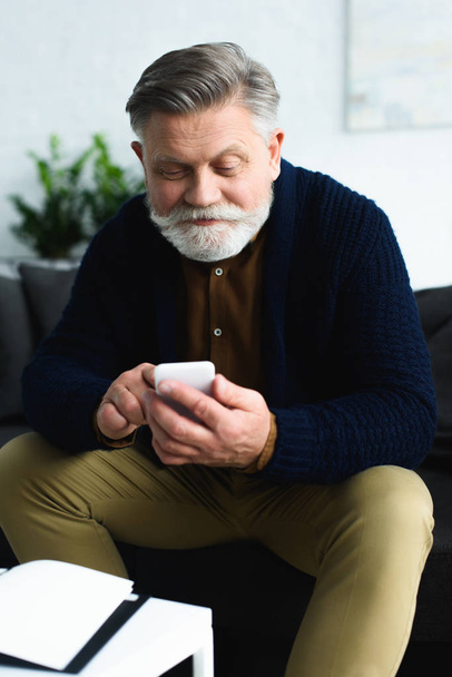 handsome smiling senior man using smartphone while sitting at home   - Foto, Bild