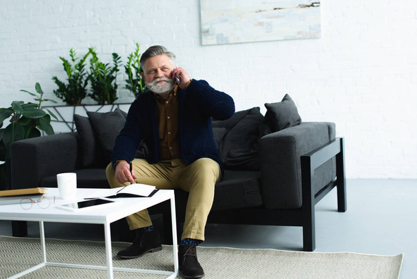 smiling bearded senior man sitting on sofa and talking by smartphone - Fotografie, Obrázek