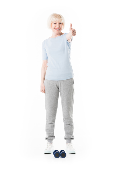Senior sportswoman showing thumb up gesture and dumbbells on floor isolated on white - Photo, Image